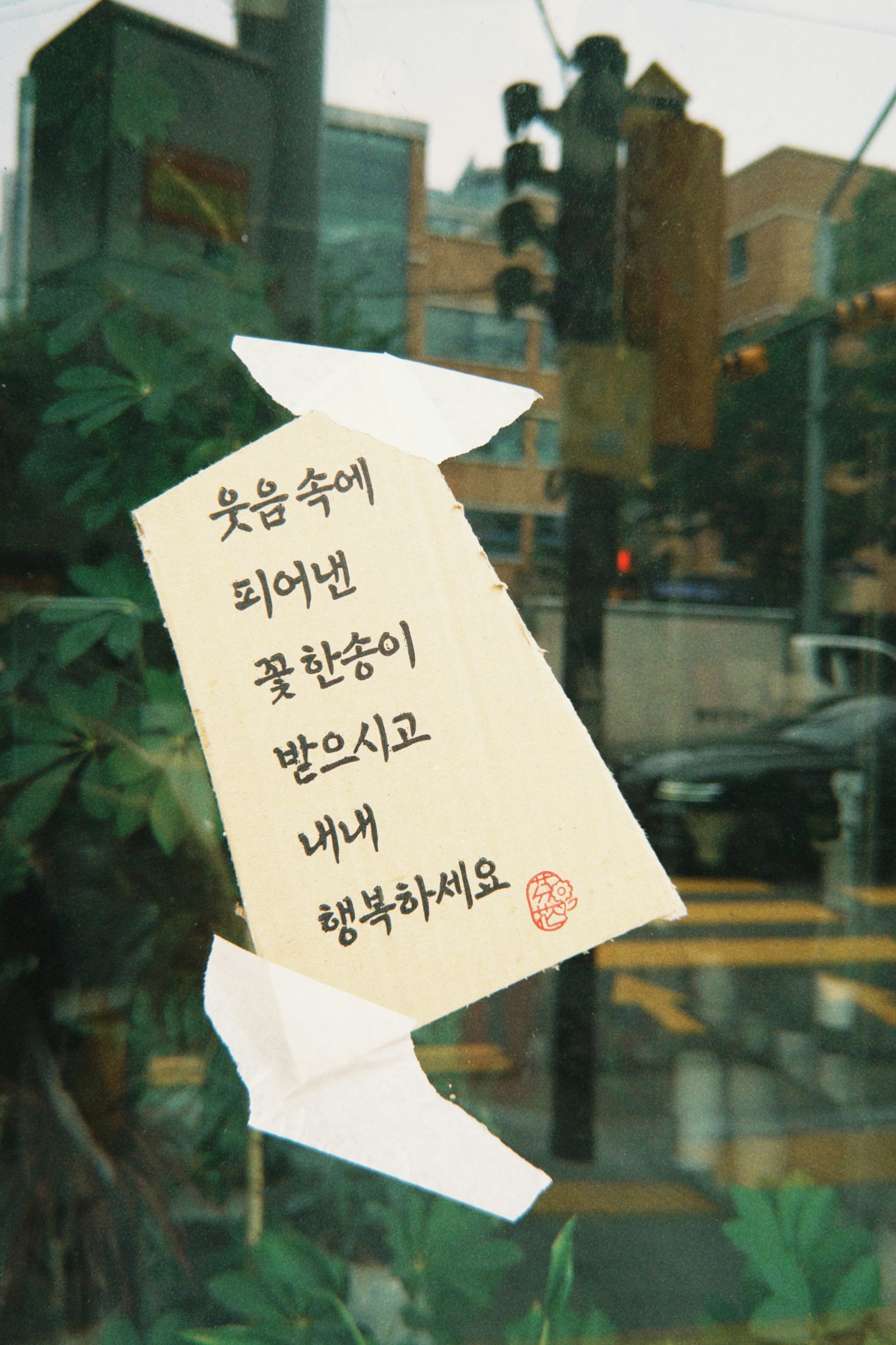 A handwritten note in Korean taped to a window, with a Korean street and a traffic light reflected in the glass behind it.