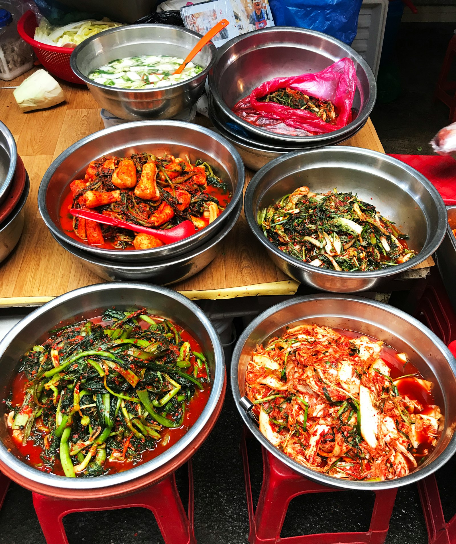 Stainless steel bowls of kimchi at a market stall, stacked on red plastic stools and a wooden board, holding several kinds: spring onion, young radish, cabbage and a watery cucumber one.