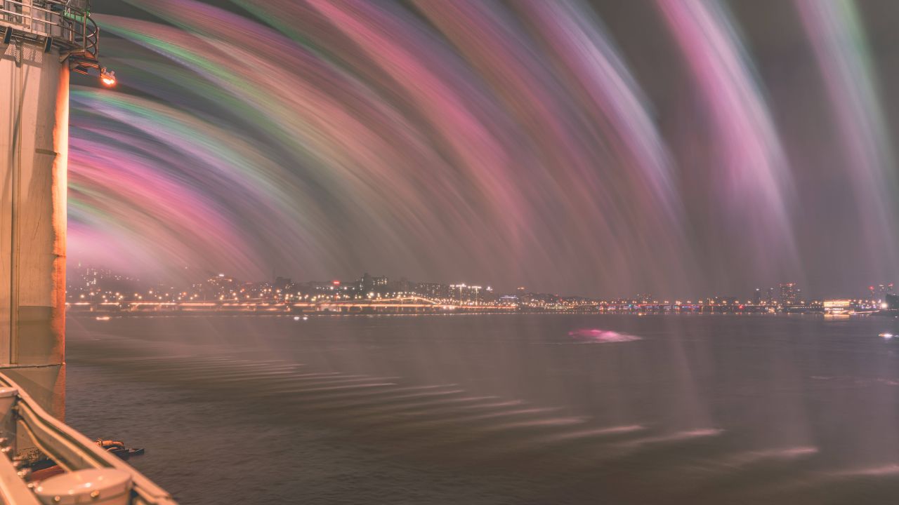 The Banpo Bridge Moonlight Rainbow Fountain at night, arcs of lit water in pink and green falling into the dark river.