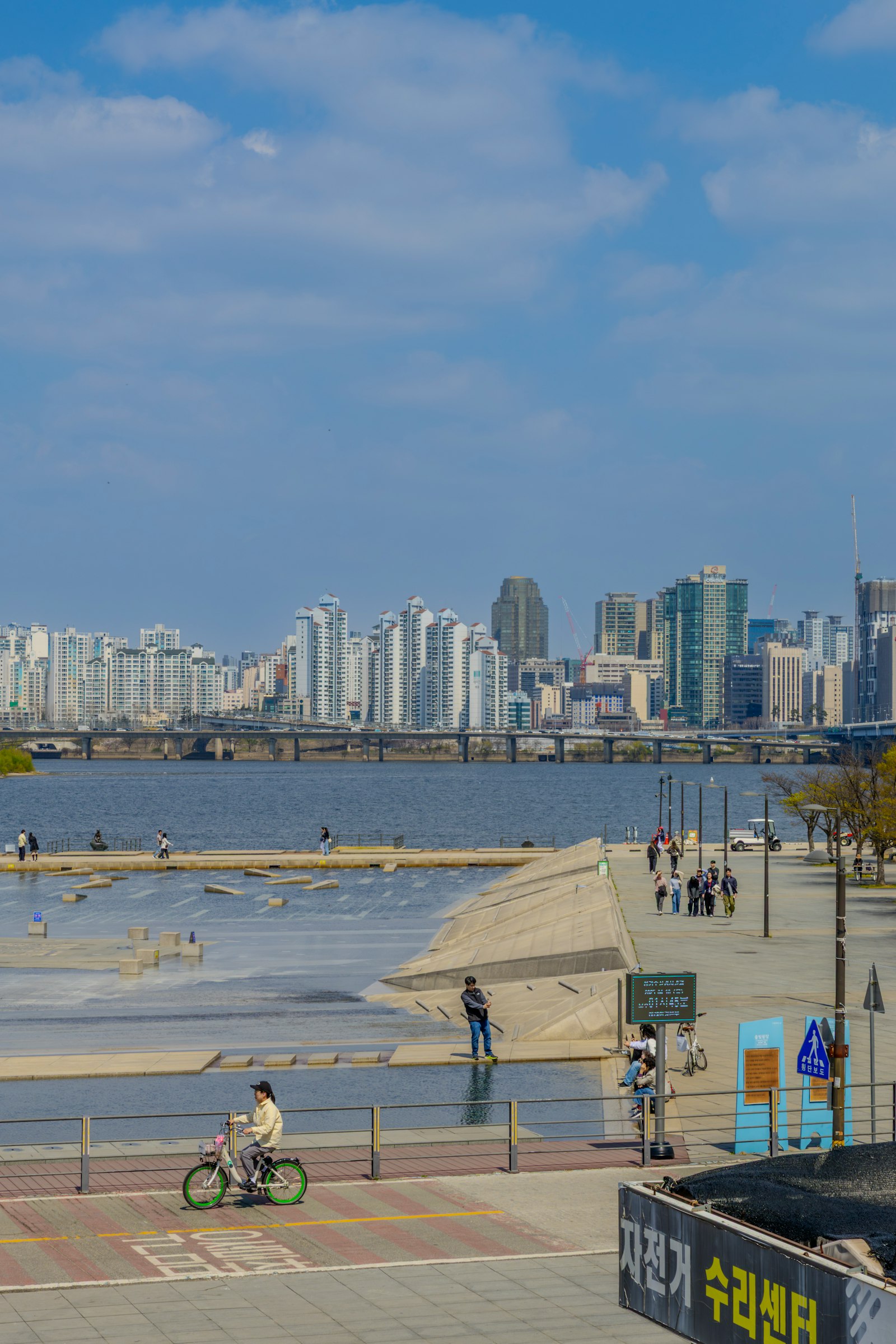 Yeouido Hangang Park on a clear spring day: a paved riverside plaza and shallow water feature, the Han River and a road bridge behind, apartment towers along the far bank.