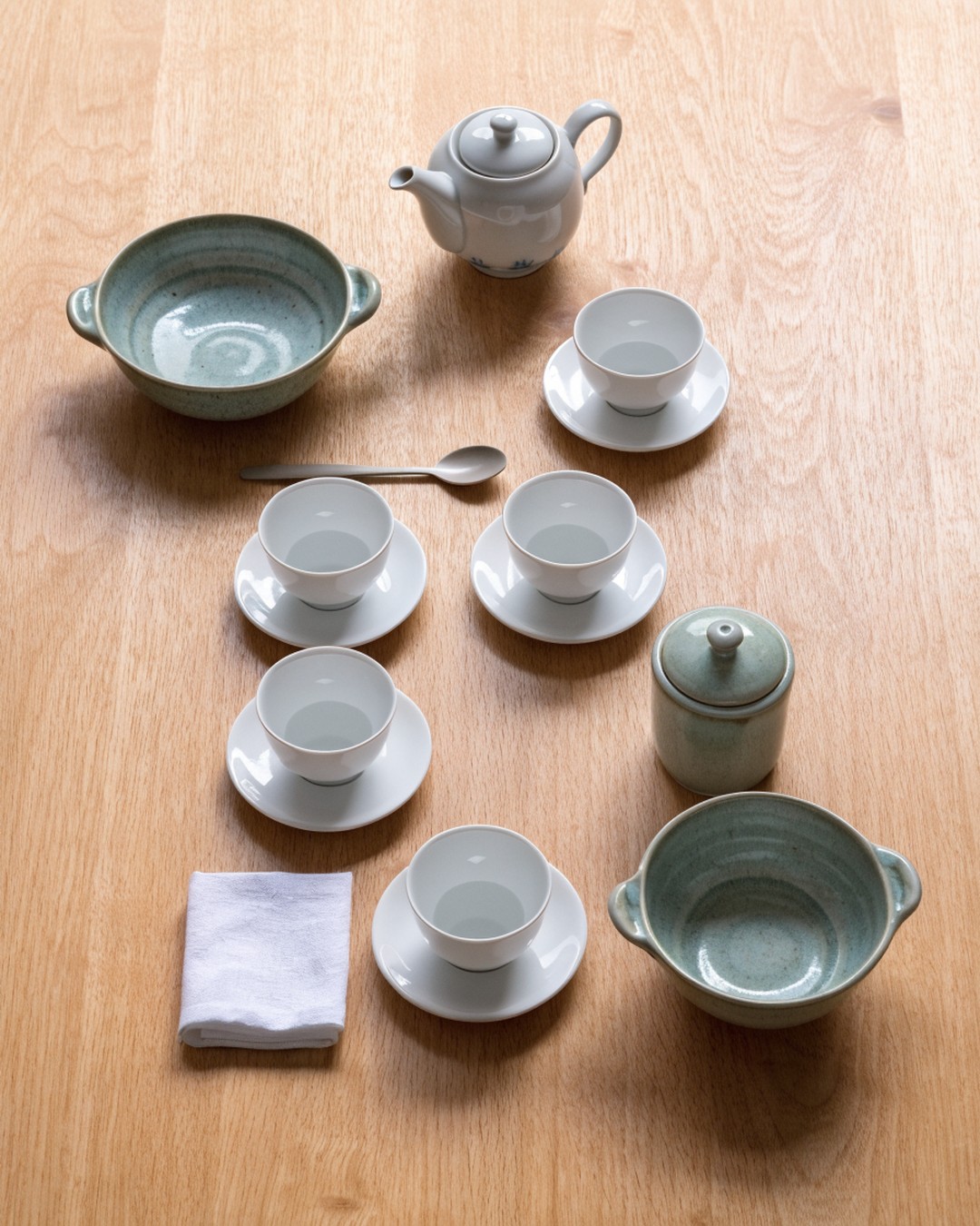 Generated image of a Korean tea set laid out on a plain wooden table: a teapot, white porcelain cups on saucers, a wide shallow bowl, a deeper bowl, a tea spoon and a folded cloth.