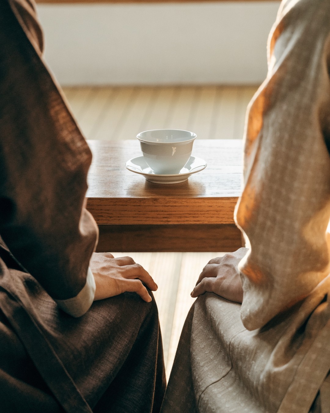 Generated image of a single white porcelain tea cup on a saucer on a wooden table between two seated people, seen from the shoulders down, faces outside the frame.